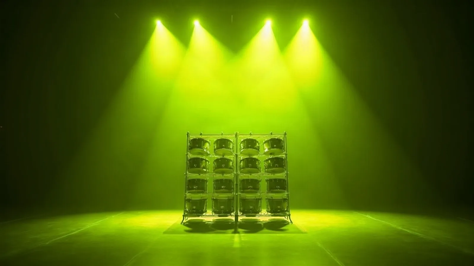 Sixteen drums held in a four-by-four steel rack, alone in a dark room under four hard lime stage lanterns, casting a row of shadows across the lit floor